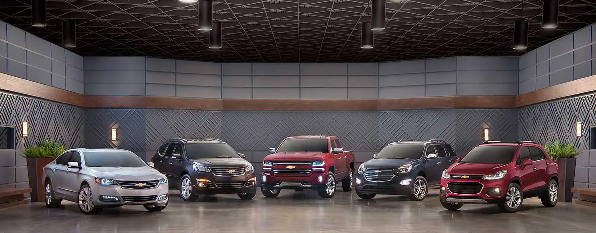 Five Chevrolet vehicles—silver Impala, dark gray Traverse, red Silverado, blue Equinox, and red Trax—in a modern showroom.