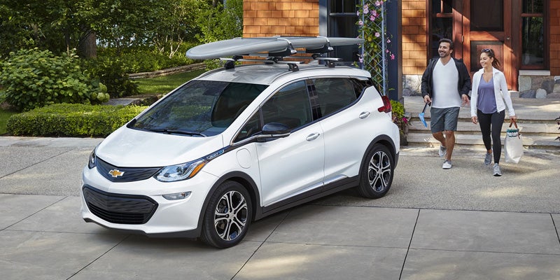 White 2020 Chevrolet Bolt EV with two surfboards on a roof rack, parked in a driveway. Two people walk towards it.