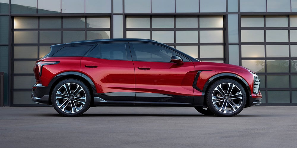 Side view of a glossy red Chevrolet Blazer EV parked against a modern building with large glass windows.