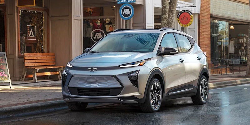 A light grey Chevy Bolt EUV electric vehicle parked on a wet city street in front of shops.