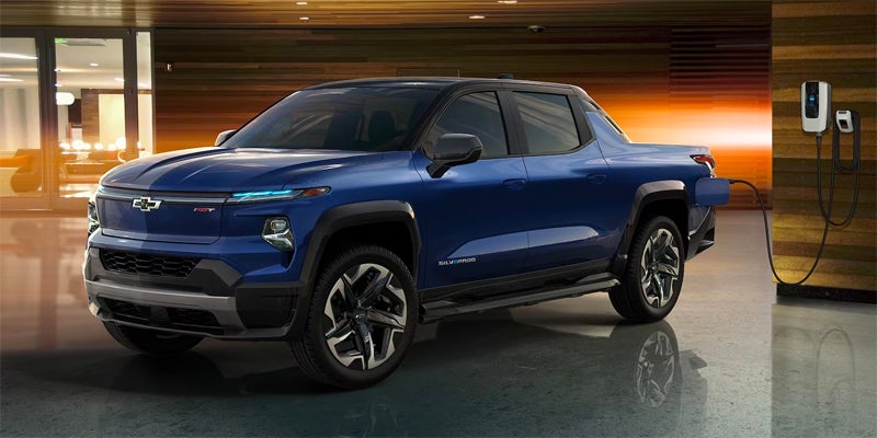 A blue Chevrolet Silverado EV pickup truck charging from a white wall-mounted charger in a modern garage.