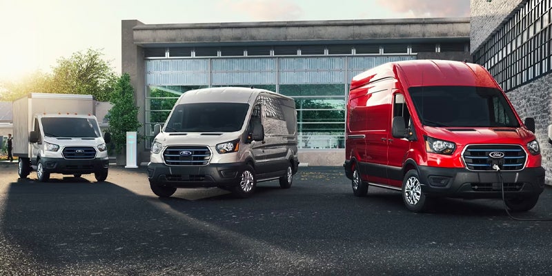 Three Ford E-Transit vans: a gray box truck, a silver cargo van, and a red cargo van being charged.