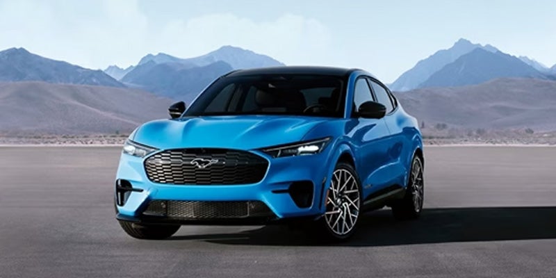 A bright blue Ford Mustang Mach-E electric SUV, angled slightly right, against a desert mountain backdrop.
