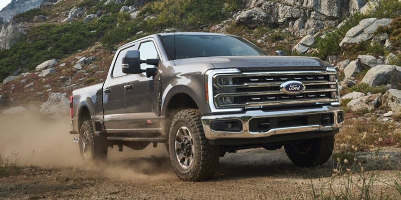 Dark gray Ford Super Duty pickup truck drives right on a dirt road, kicking up dust against a rocky hillside.