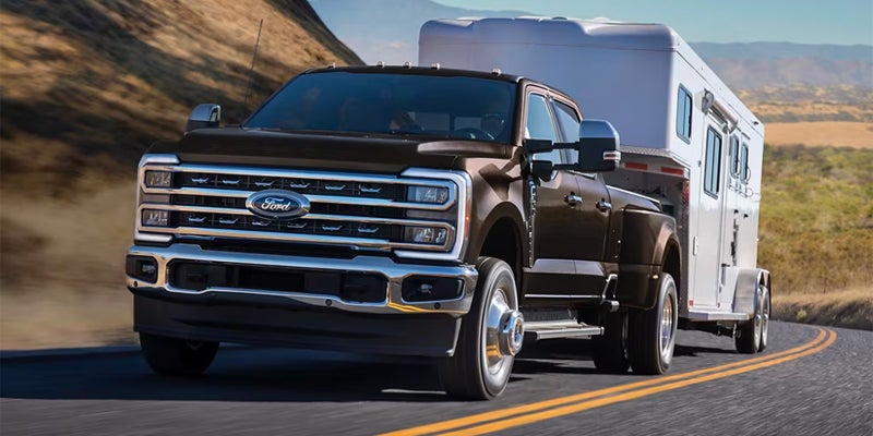 A Ford Super Duty pickup truck in dark brown tows a white trailer on a winding road with hills in the background.
