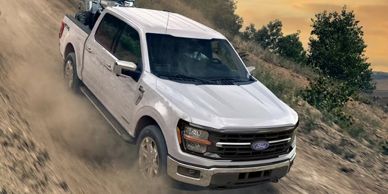 A white Ford F-150 pickup truck with an ATV in its bed drives down a dusty dirt hill at sunset.