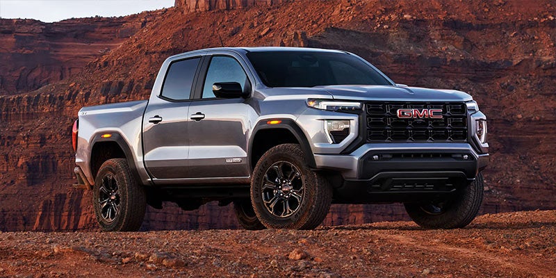 A silver-gray GMC Canyon pickup truck with black wheels and aggressive styling sits on a dirt road in a desert landscape.