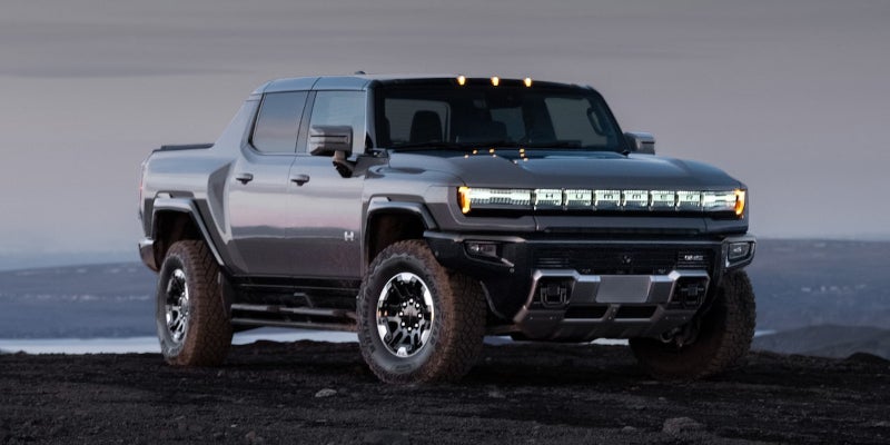 A gray Hummer EV pickup truck with illuminated headlights and roof lights, parked on a dark, rocky terrain.