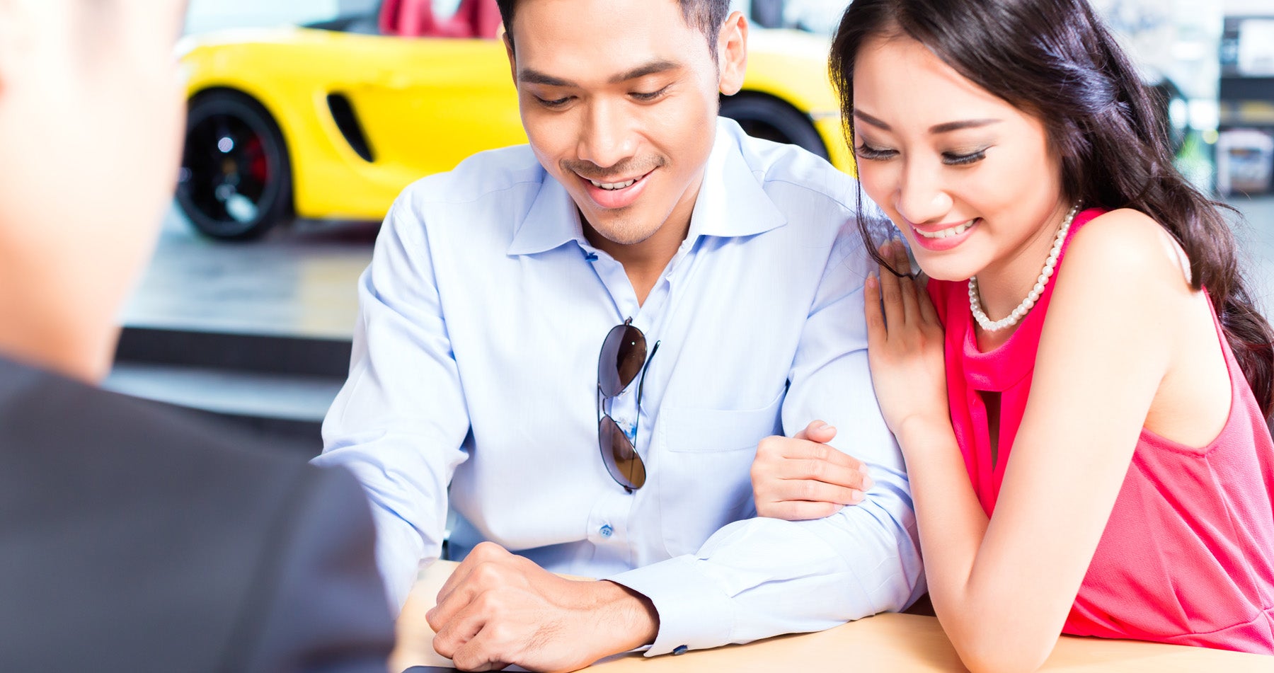 A smiling couple sits at a desk in a car showroom, presumably discussing a purchase with a salesperson.
