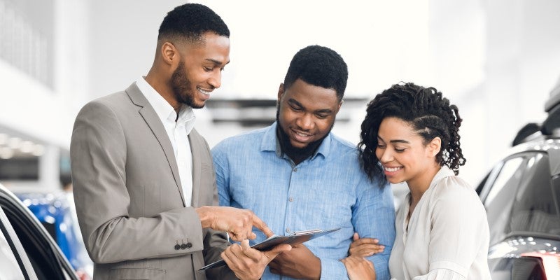 A car salesman in a suit reviews documents on a clipboard with a happy couple in a dealership.