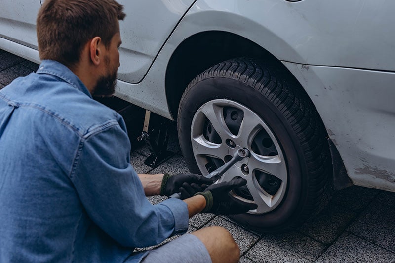 Man in denim shirt and gloves loosening lug nuts on car tire with a lug wrench, car jacked up.