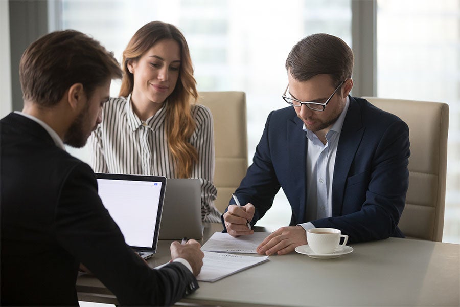 Three business professionals review documents at a conference table with a laptop and coffee.