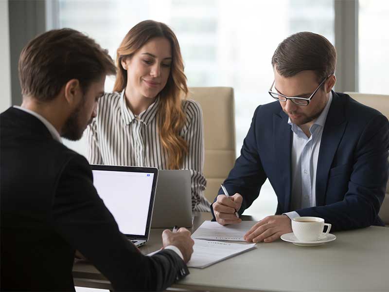 Three businesspeople review documents and a laptop in a brightly lit office setting.