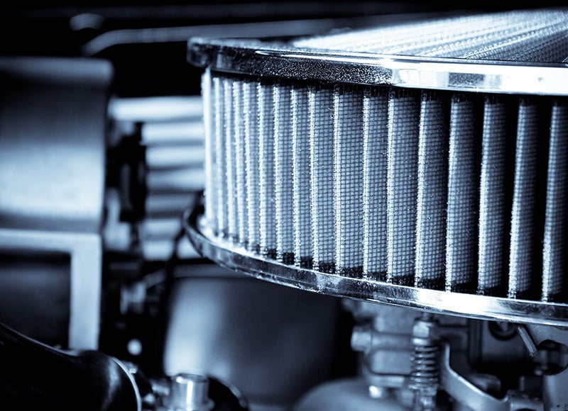 Close-up, high-contrast, monochrome shot of a car's engine with a round, chrome-ringed air filter in the foreground.