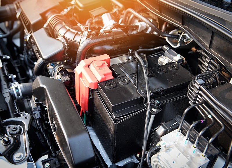 A car engine bay with a black battery and red terminal cover, various hoses, and components.
