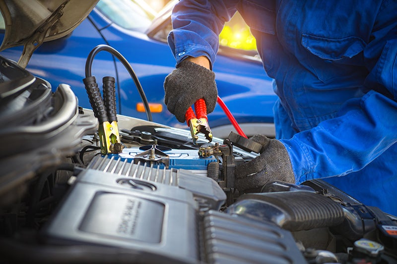 A person in blue overalls and gloves connecting red and black jumper cables to a car battery.