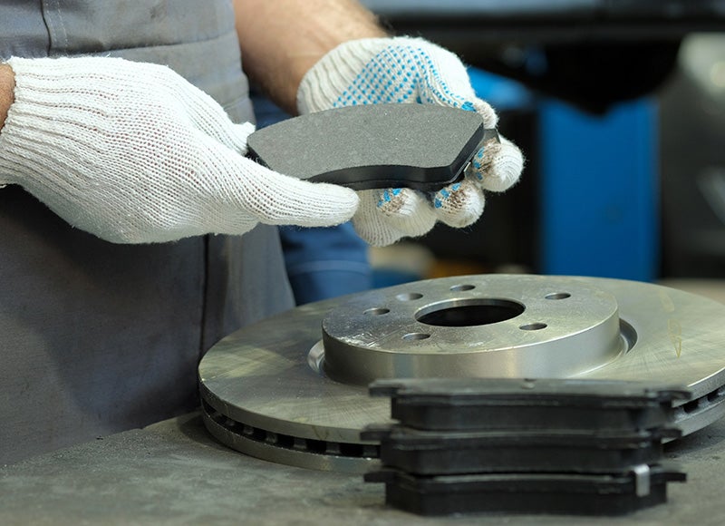 Gloved hands hold a new brake pad over a new brake rotor, with more pads stacked nearby.