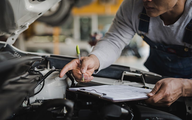 Mechanic in overalls pointing at a car engine while holding a pen and clipboard to check off a list.