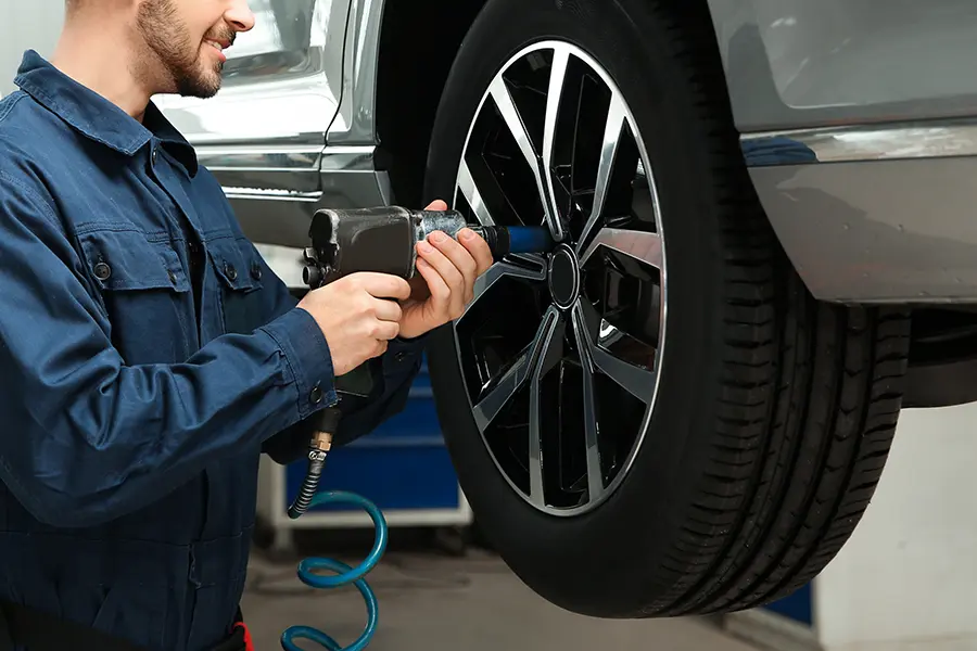 Smiling mechanic in blue overalls uses an impact wrench to change a tire on a raised silver car.