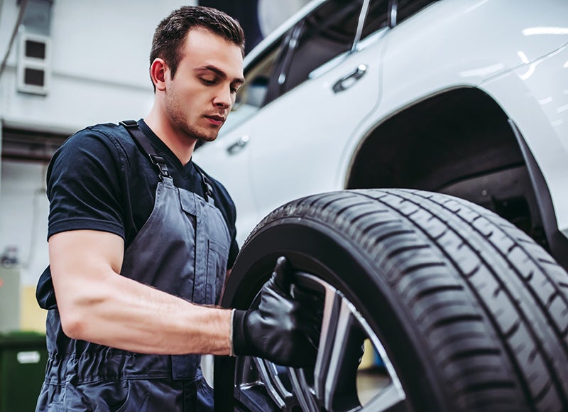 A male auto mechanic in gray overalls and gloves mounts a tire in front of a white SUV.