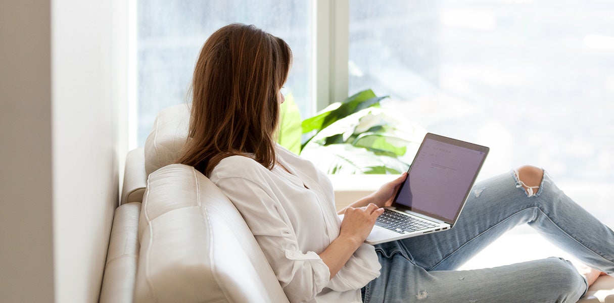 Woman with long brown hair, sitting on a couch near a window, typing on a laptop.
