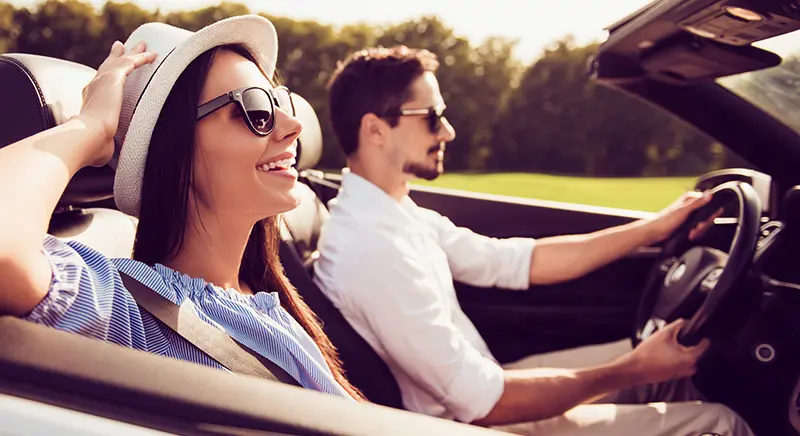 Smiling woman in sunglasses and hat riding in a convertible with a man driving.